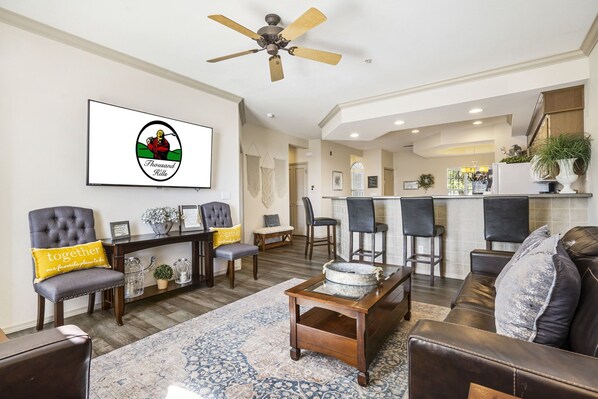 A spacious living room with a large TV, comfortable seating, a coffee table, and a kitchen area in the background. - Walk-in Condo • Golf Views •  Sunroom   • Show Discounts (Branson)