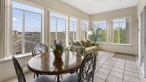 Uma sala iluminada pelo sol com uma mesa de jantar, cadeiras, um sofá e grandes janelas com vista para o bairro.