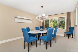 Een eetkamer met een ronde tafel en zes blauwe stoelen, een kroonluchter en een raam met gordijnen.