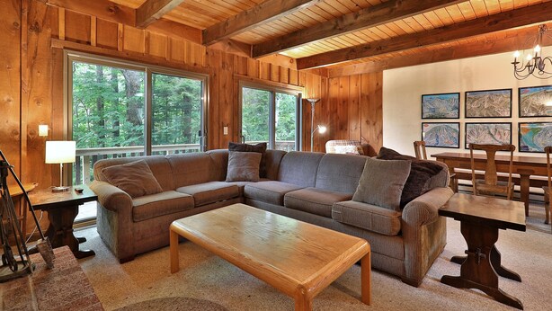 A cozy cabin interior with a large sectional sofa, a wooden coffee table, and a dining table with chairs. Large windows offer views of the surrounding forest. - Hemlock Ridge 2B: Charming two floor condo in the heart of Killington. Perfect! (Killington)