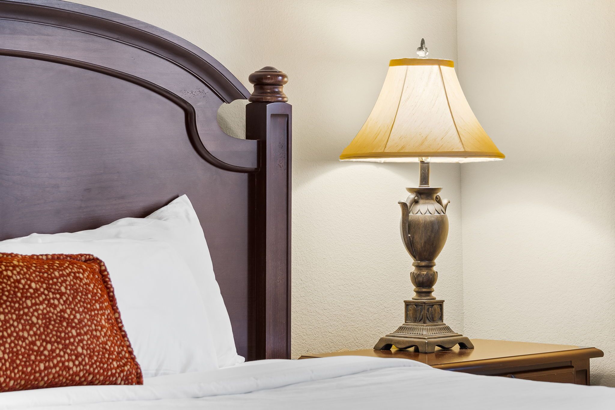 Un lit bien fait, avec un oreiller blanc et une couverture à motifs, une table de chevet en bois avec une lampe, et une tête de lit en bois foncé.