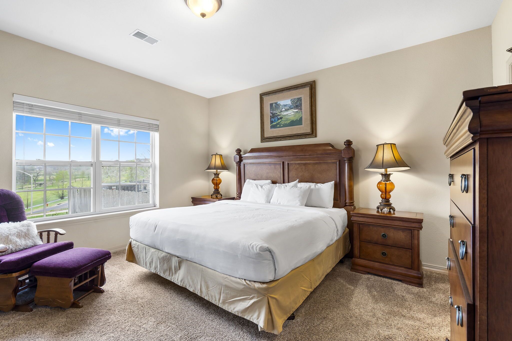 A bedroom with a large bed, two nightstands, a dresser, a chair, and a window with a view.