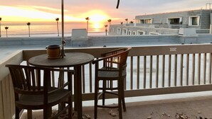 A balcony with a table and chairs, overlooking a beach at sunset.