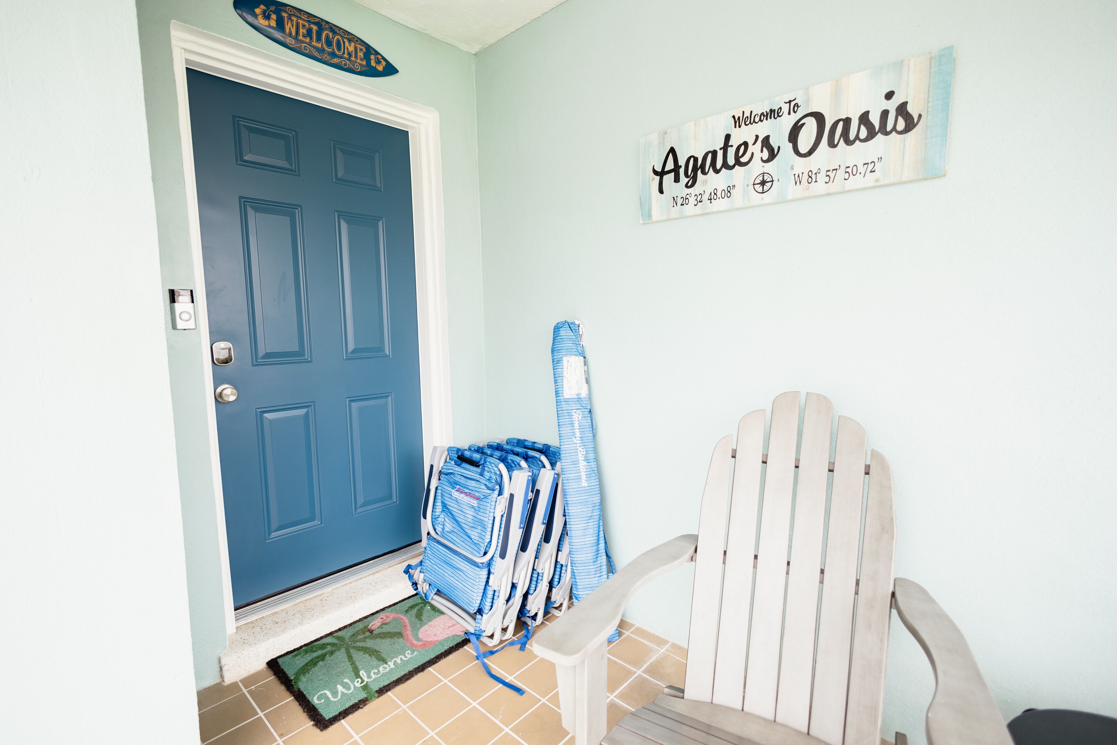 A blue door with a welcome sign, a beach chair, and a mat with a palm tree design.