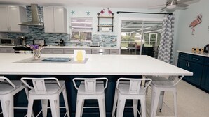Een moderne keuken met witte kasten, een groot kookeiland met barstoelen, een plafondventilator en decoratieve flamingo’s aan de muur.