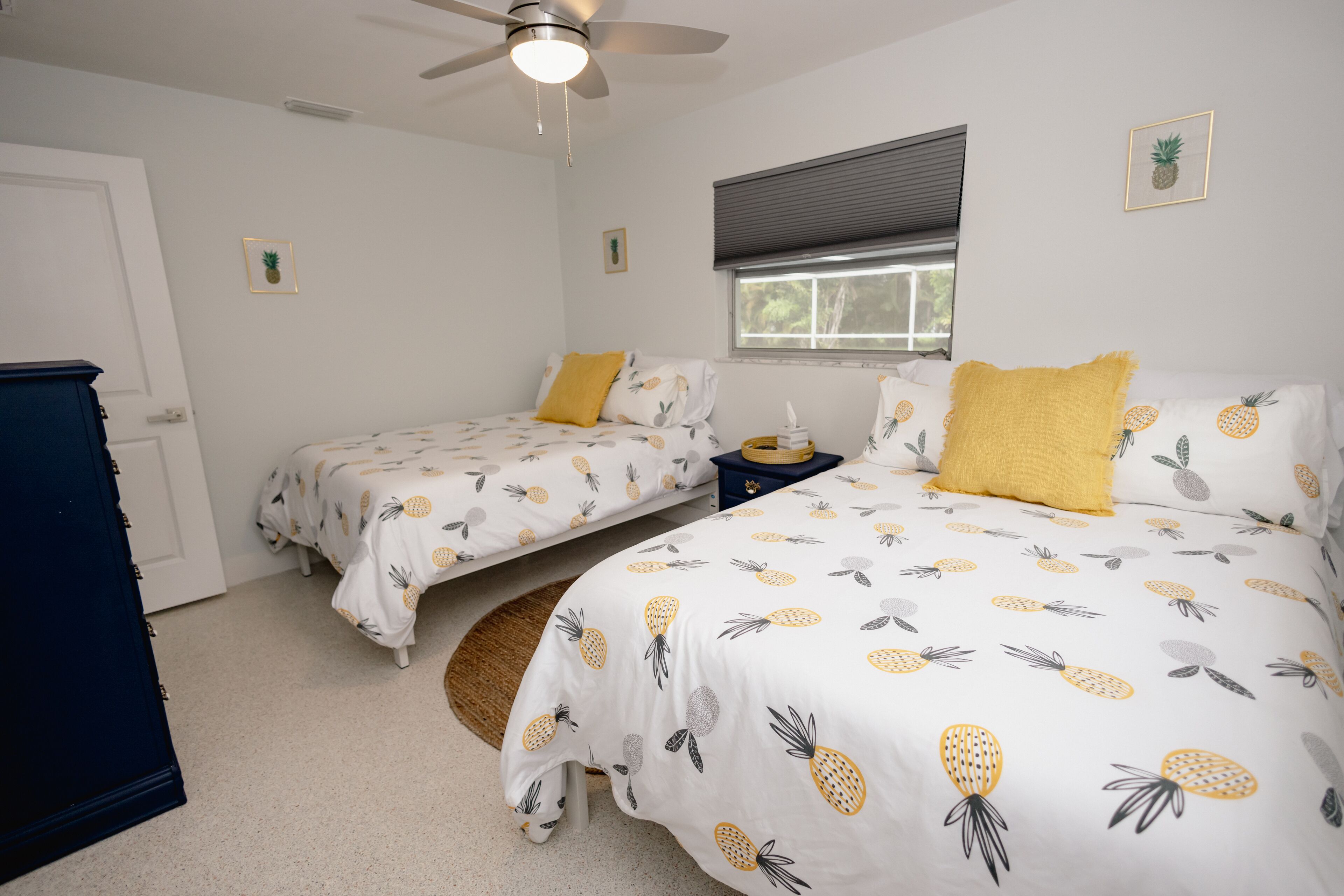 A bedroom with two beds, each with a pineapple-themed bedspread, a ceiling fan, and a window with blinds.