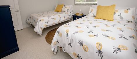 A bedroom with two beds, each with a pineapple-themed bedspread, a ceiling fan, and a window with blinds.