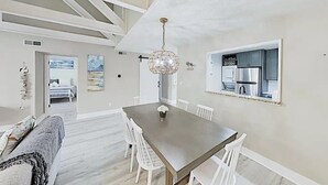 Un espace repas avec une longue table, des chaises blanches, un lustre et une cuisine visible à travers une porte ouverte.