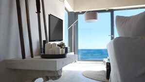 Habitació moderna amb gran finestra que dona al mar, televisió penjada a la paret i una tauleta de nit amb espelmes.