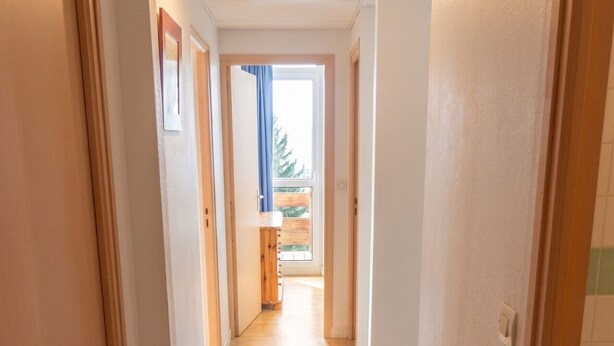 A hallway with wooden flooring and white walls, leading to a room with a window and a view of greenery outside. - Homerez - Apartment in Allevard (Allevard)