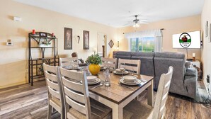 Un coin repas avec une table en bois dressée pour quatre personnes, entourée de chaises. Un présentoir en métal décoratif, orné de bougies et d’une plante en pot, est accroché au mur. Un canapé gris et un téléviseur avec un logo se trouvent à l’arrière-plan.