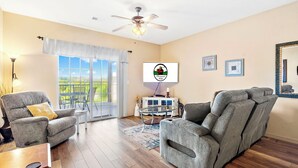 Un salon avec deux fauteuils inclinables gris, un ventilateur de plafond, une grande fenêtre offrant une belle vue et un meuble blanc agrémenté d’objets décoratifs.