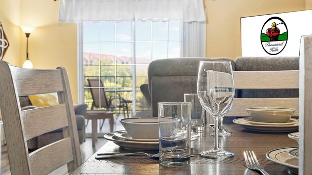 Une table à manger dressée avec des assiettes, des bols, des verres et des couverts, donnant sur l’extérieur par une fenêtre.