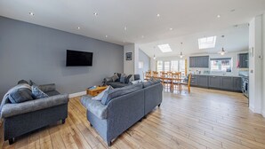 Een ruime woonkamer met een grijze zithoek, houten vloeren, een flatscreen televisie en een keuken op de achtergrond.