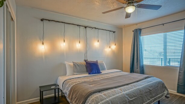 Ein Schlafzimmer mit einem Bett, einem Deckenventilator, einer Lichterkette, einem Nachttisch und einem Fenster mit Vorhängen.