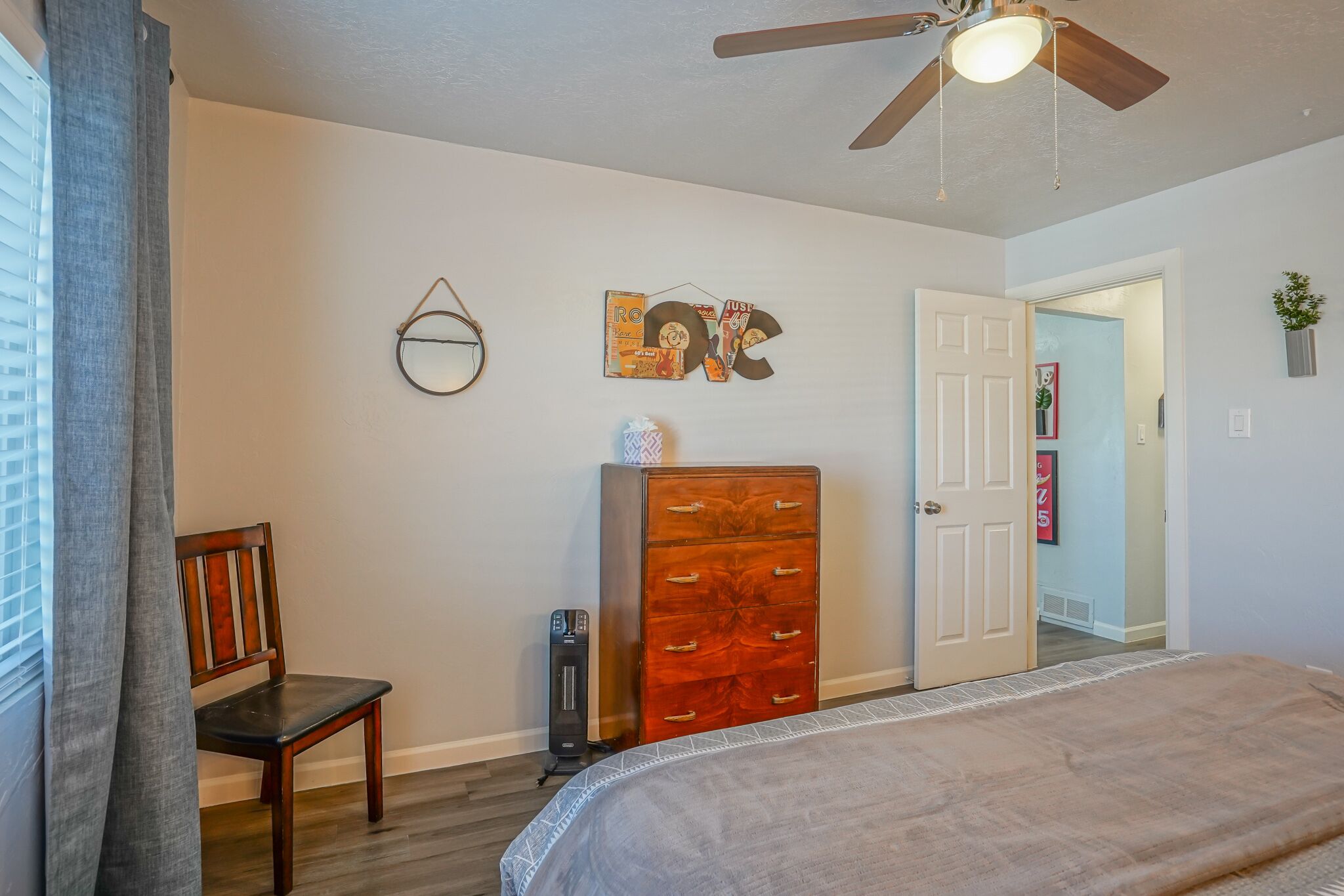 A bedroom with a bed, a dresser, a chair, a ceiling fan, and a door leading to another room.