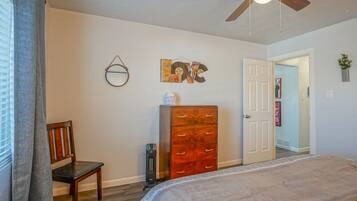 A bedroom with a bed, a dresser, a chair, a ceiling fan, and a door leading to another room.