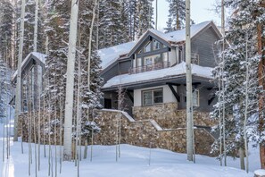 A large, multi-story house with a stone foundation and a wooden upper level, surrounded by tall, snow-covered trees.