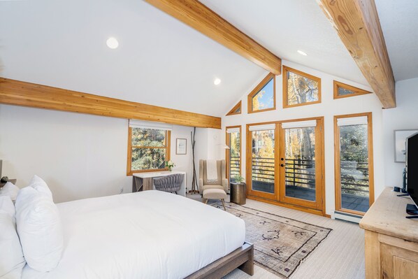 A bedroom with a large bed, wooden ceiling beams, a desk, a chair, and a TV.