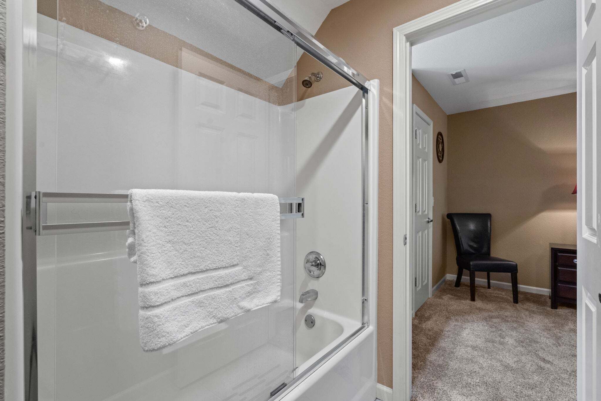 Une douche en verre avec une serviette blanche, une porte et une chaise, dans une pièce aux murs et au tapis de couleur beige.