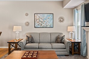 A living room with a grey sofa, two side tables with lamps, a coffee table with a board game, and a framed floral painting on the wall.