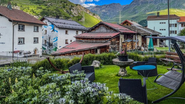 A traditional Swiss village with a mountain backdrop, featuring a mix of wooden and stone buildings, a flag, and a well-maintained garden with outdoor seating.