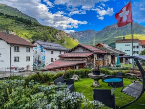 A traditional Swiss village with a mountain backdrop, featuring a mix of wooden and stone buildings, a flag, and a well-maintained garden with outdoor seating. - Vacation apartment with a panoramic view of the Swiss mountains (Surses)