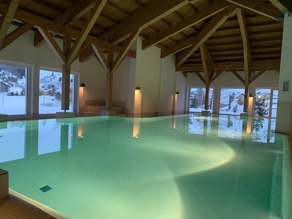 An indoor swimming pool with wooden beams, large windows, and a view of a snowy landscape.