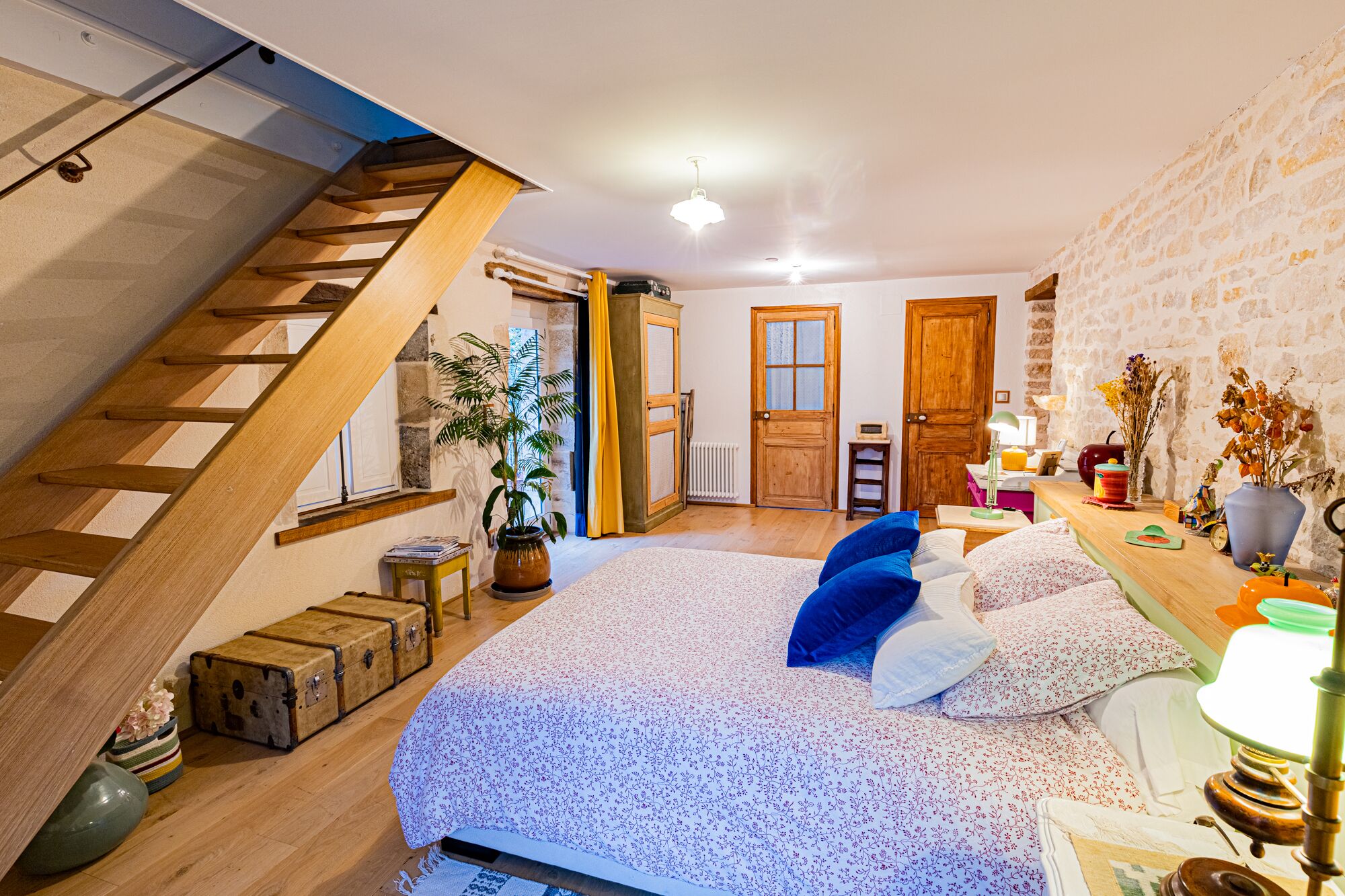 A bedroom with a wooden staircase, a bed with floral bedding, a small table with a lamp, and a brick wall.