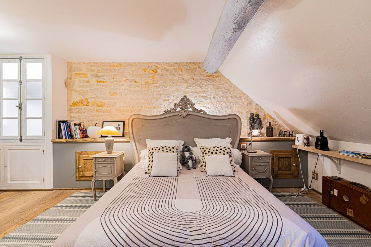A bedroom with a stone wall, a wooden bed, a nightstand, a lamp, and a small shelf with books and a statue.
