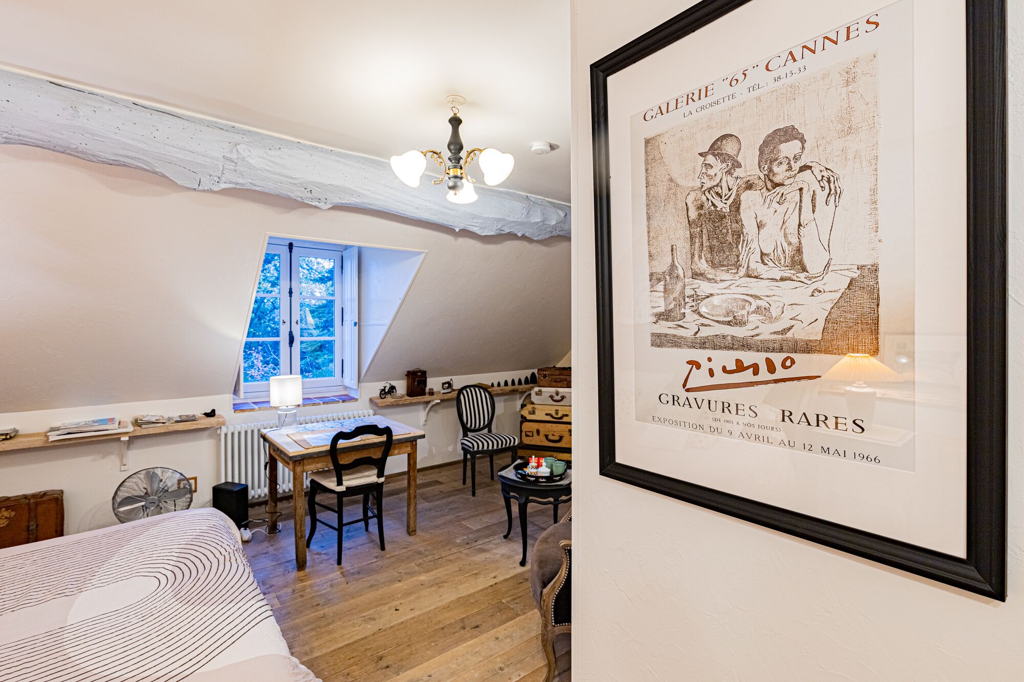 A cozy room with a sloped ceiling, a wooden table with chairs, a window with a view, and a framed poster on the wall.