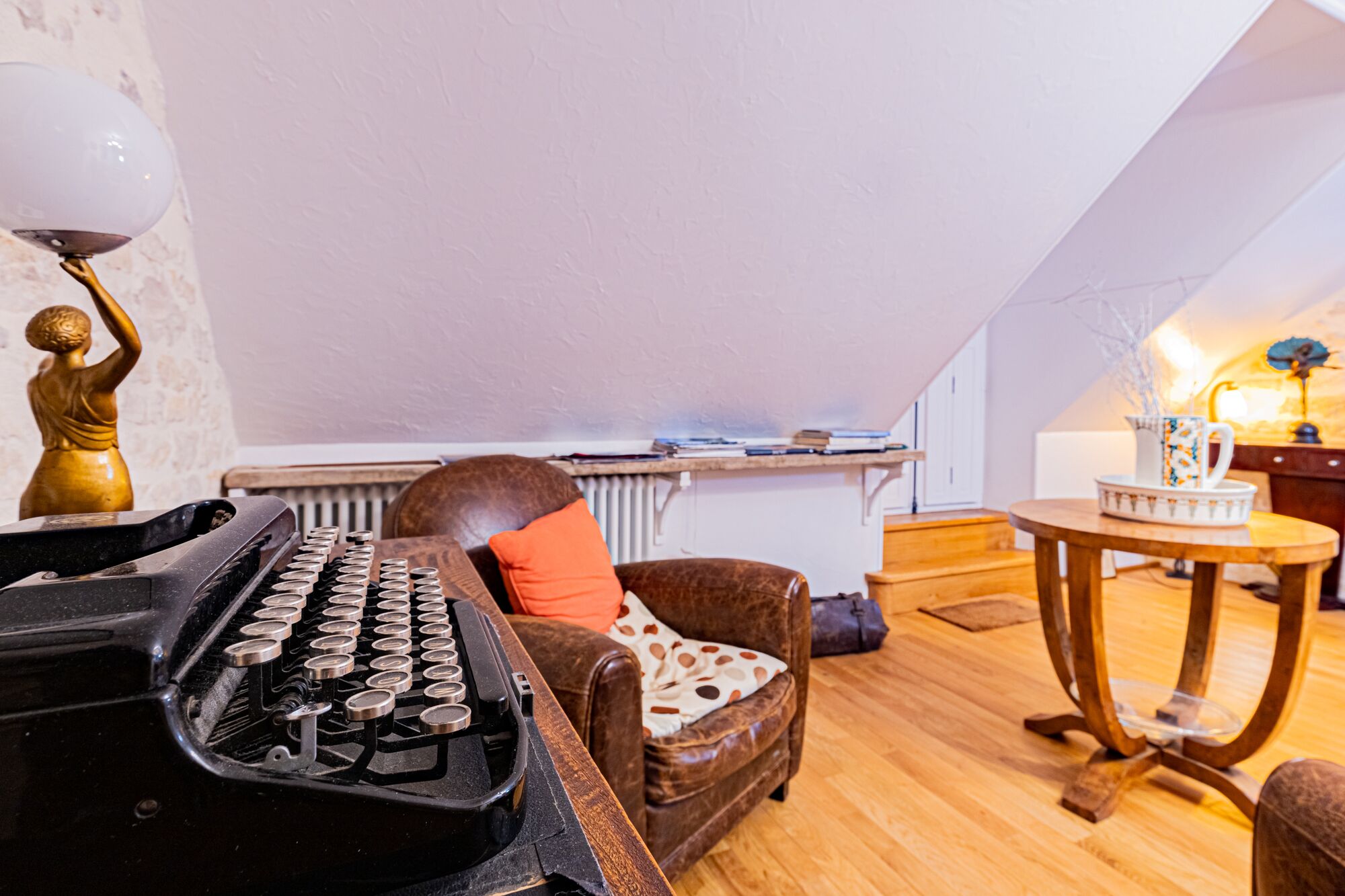 Un salon chaleureux avec une machine à écrire ancienne, un fauteuil en cuir marron, une petite table ronde en bois et un radiateur en arrière-plan.
