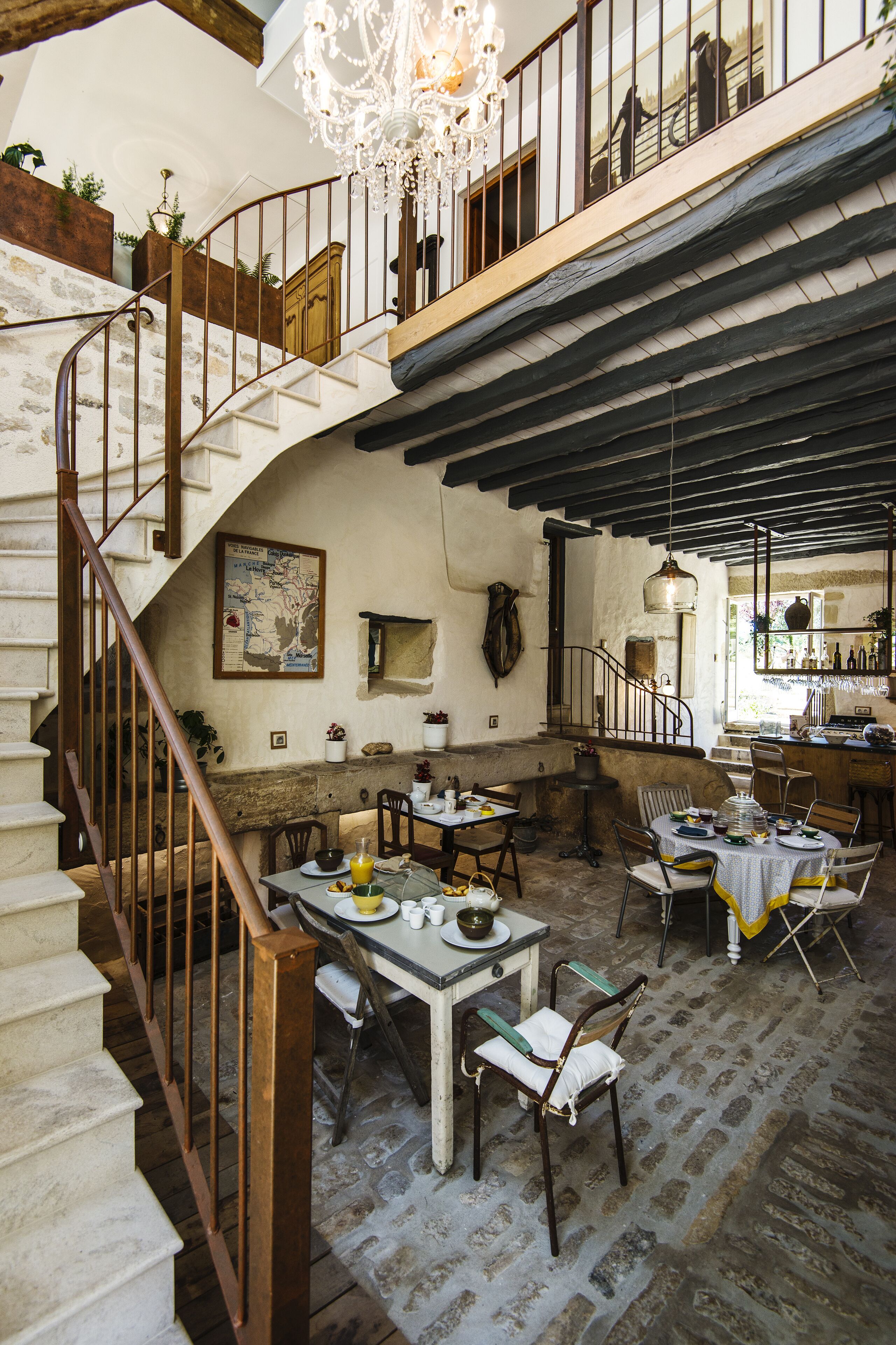 A rustic interior with a spiral staircase, stone walls, wooden beams, and a dining area with tables and chairs.