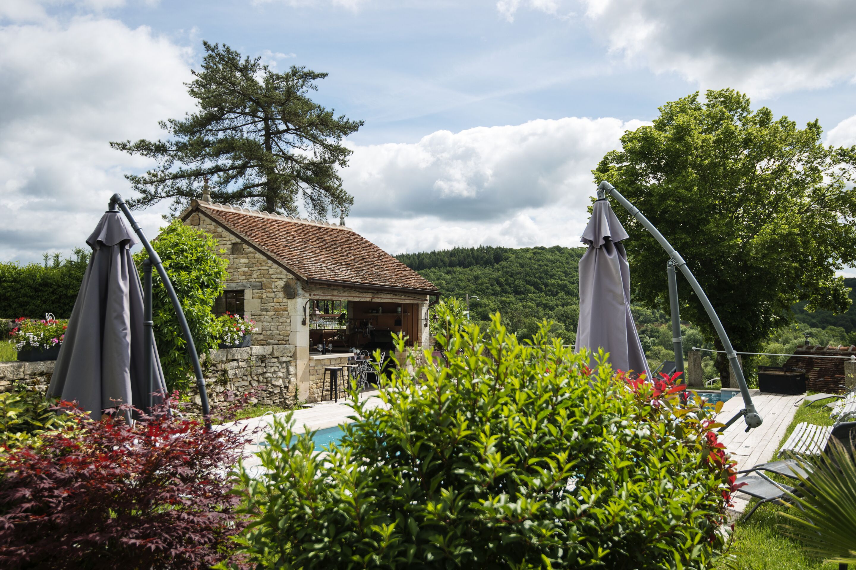 A stone building with a tiled roof, surrounded by lush greenery and a swimming pool.