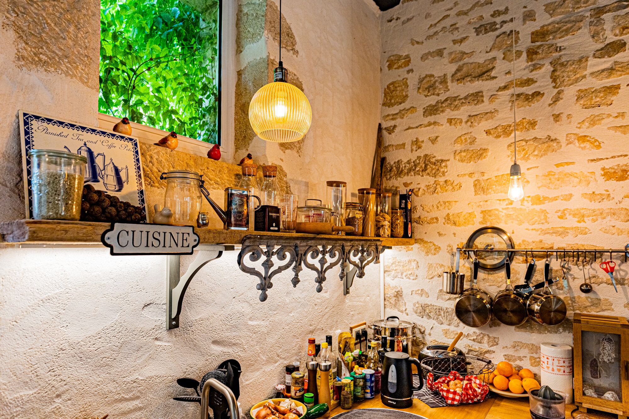 Una cocina rústica con un letrero que dice 'CUISINE', diversos utensilios de cocina y una ventana con follaje.