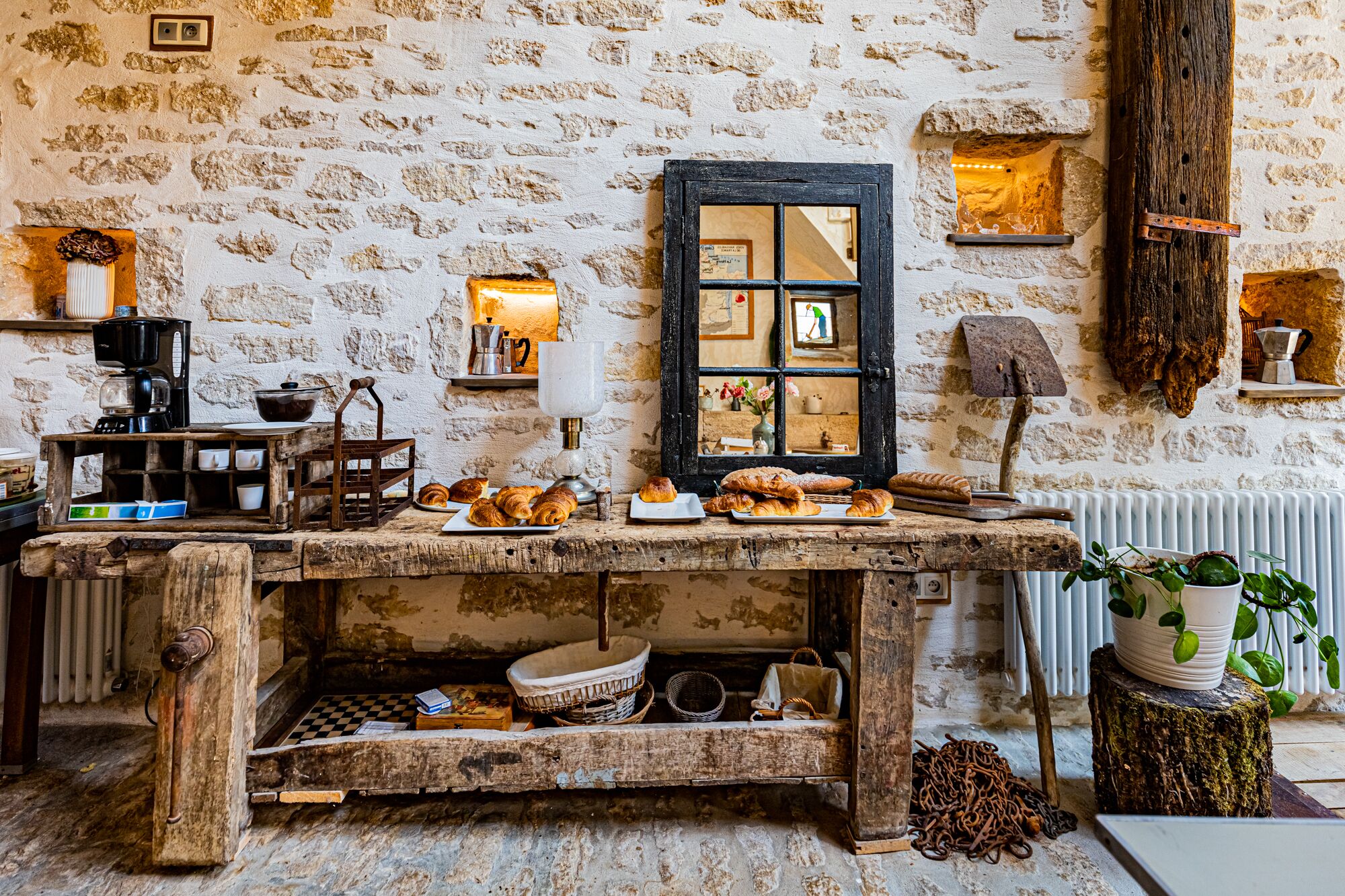 A rustic kitchen with stone walls, a wooden table with pastries, a coffee machine, and a vintage lamp.