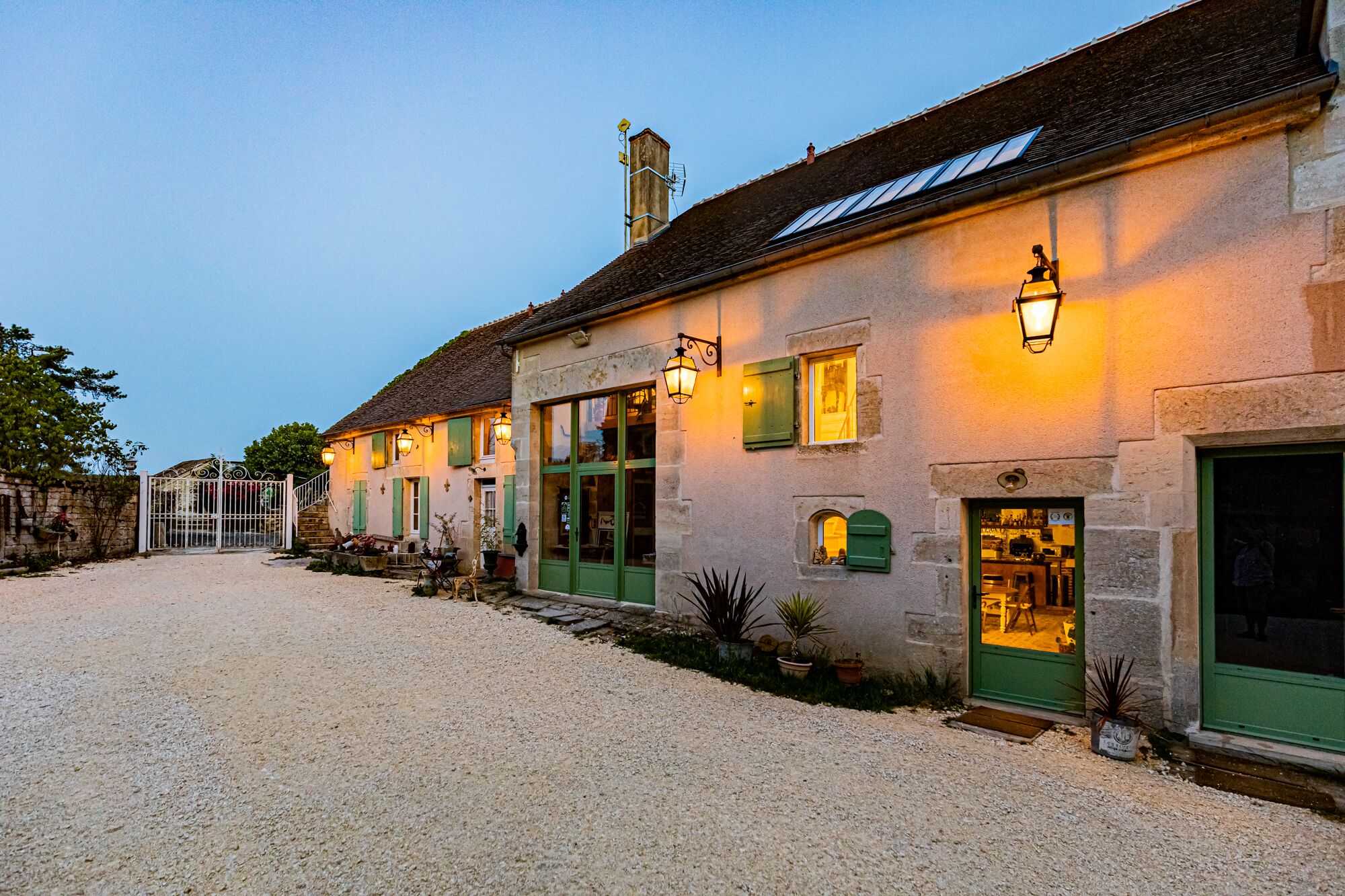 Un bâtiment doté de nombreuses portes et fenêtres, dont certaines sont agrémentées de volets verts, et une allée menant à la maison est recouverte de gravier.