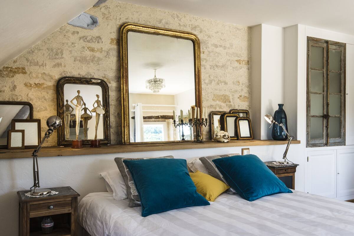 A bedroom with a large mirror, a bed with blue and yellow pillows, a nightstand, and a dresser.