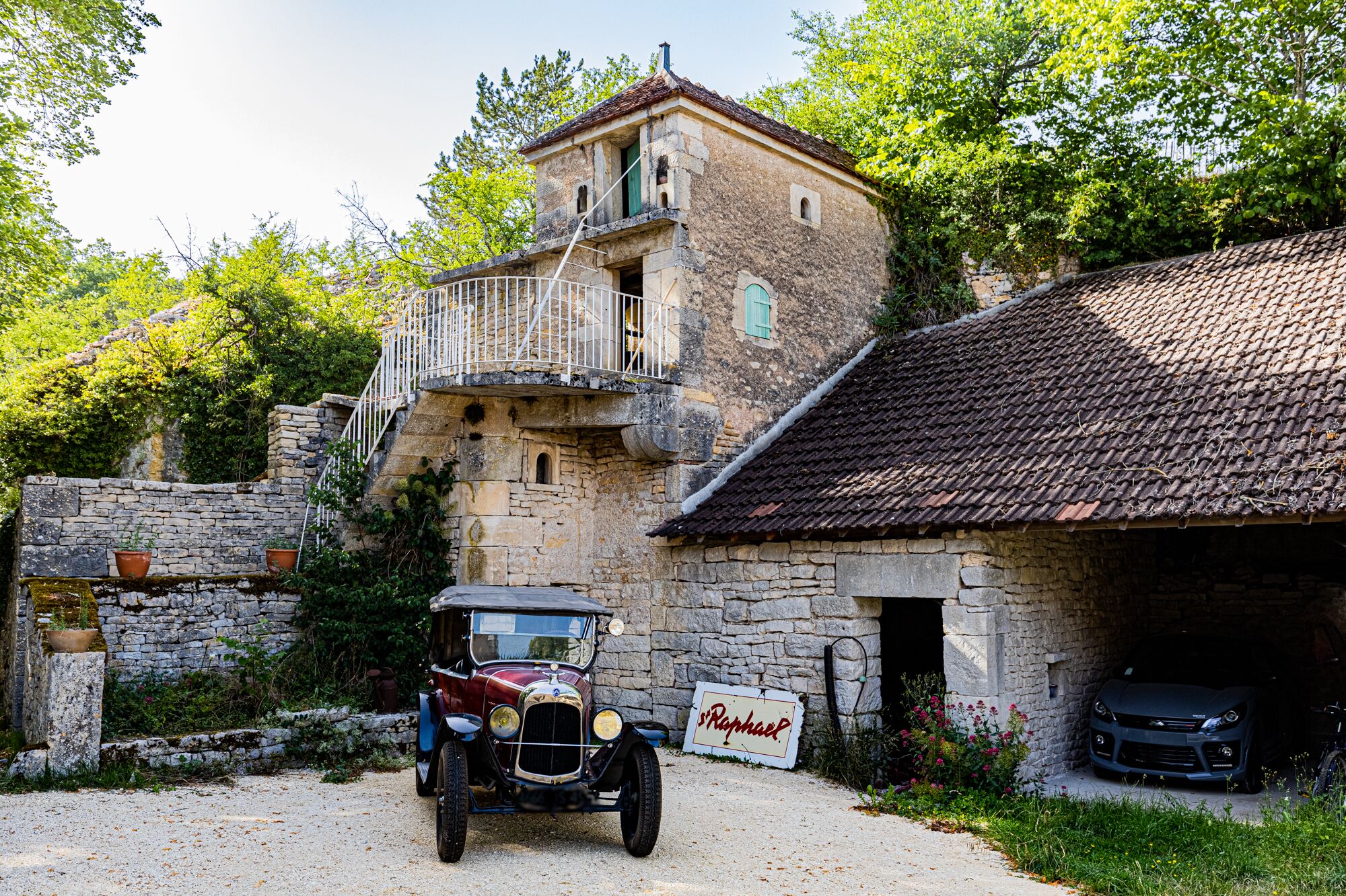 Ein Oldtimer vor einem Steinhaus, auf dessen Schild „St. Raphael“ steht.