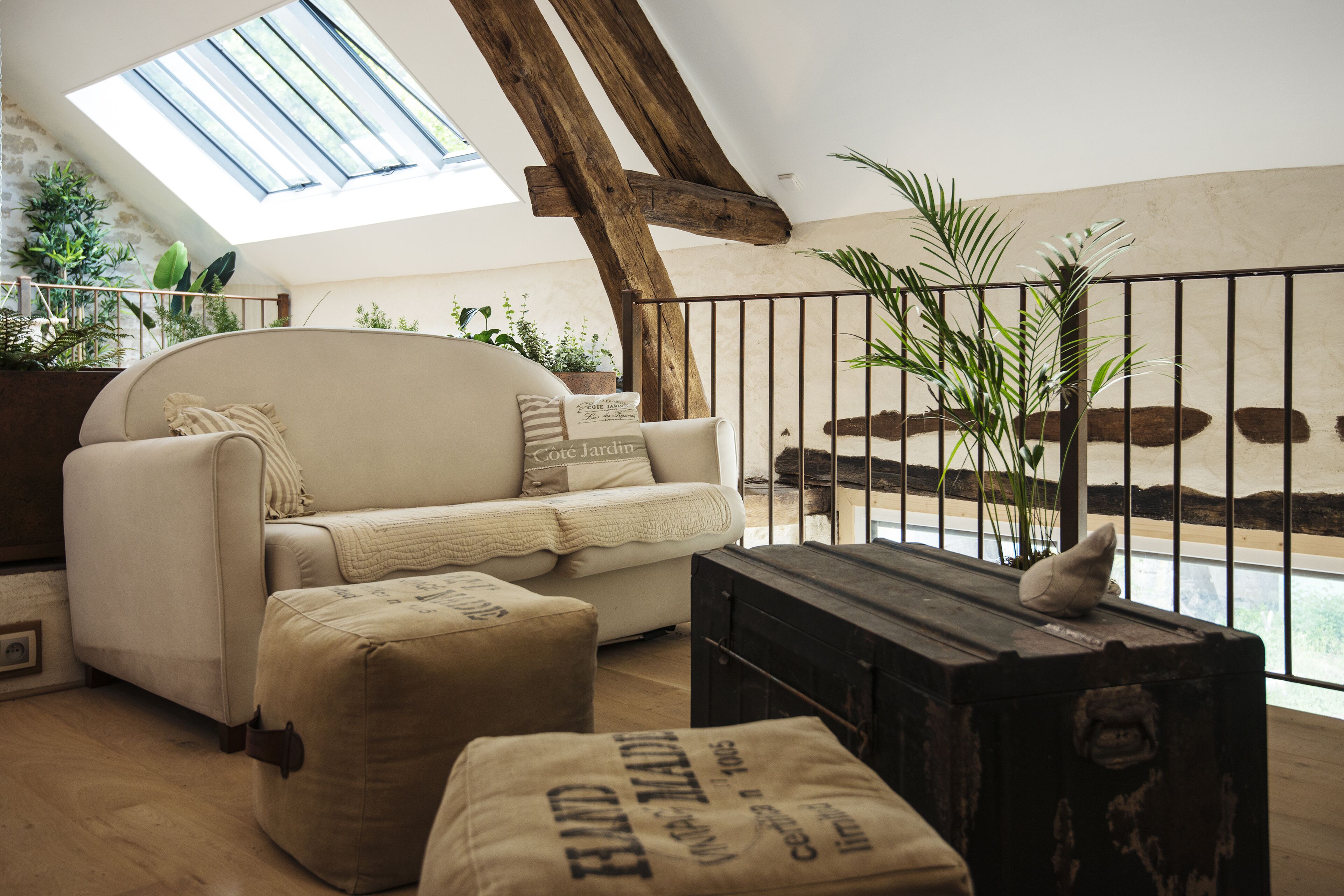 Un salon chaleureux avec un plafond haut, un canapé, des poufs, une malle en bois et une vue sur un jardin par la fenêtre.
