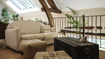 Un salon chaleureux avec un plafond haut, un canapé, des poufs, une malle en bois et une vue sur un jardin par la fenêtre.
