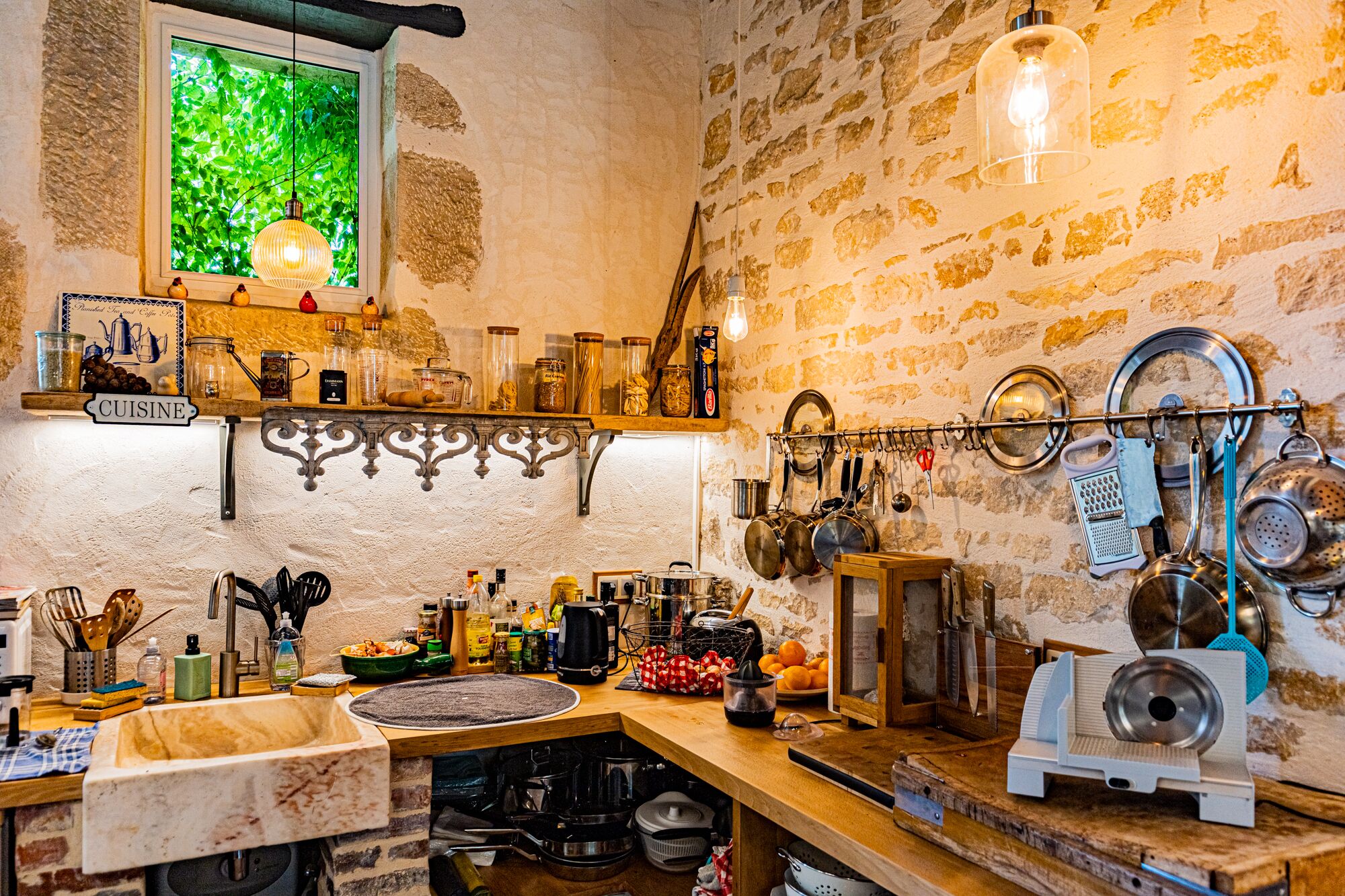 A rustic kitchen with a stone wall, a window with greenery view, a wooden countertop, and various kitchen utensils and appliances.