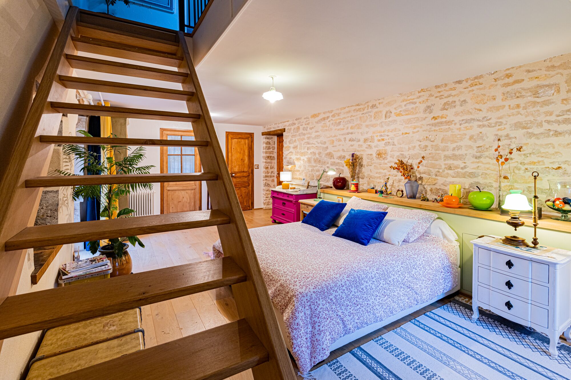 A bedroom with a wooden ladder, a bed with blue and white bedding, a nightstand with a lamp, and a stone wall.