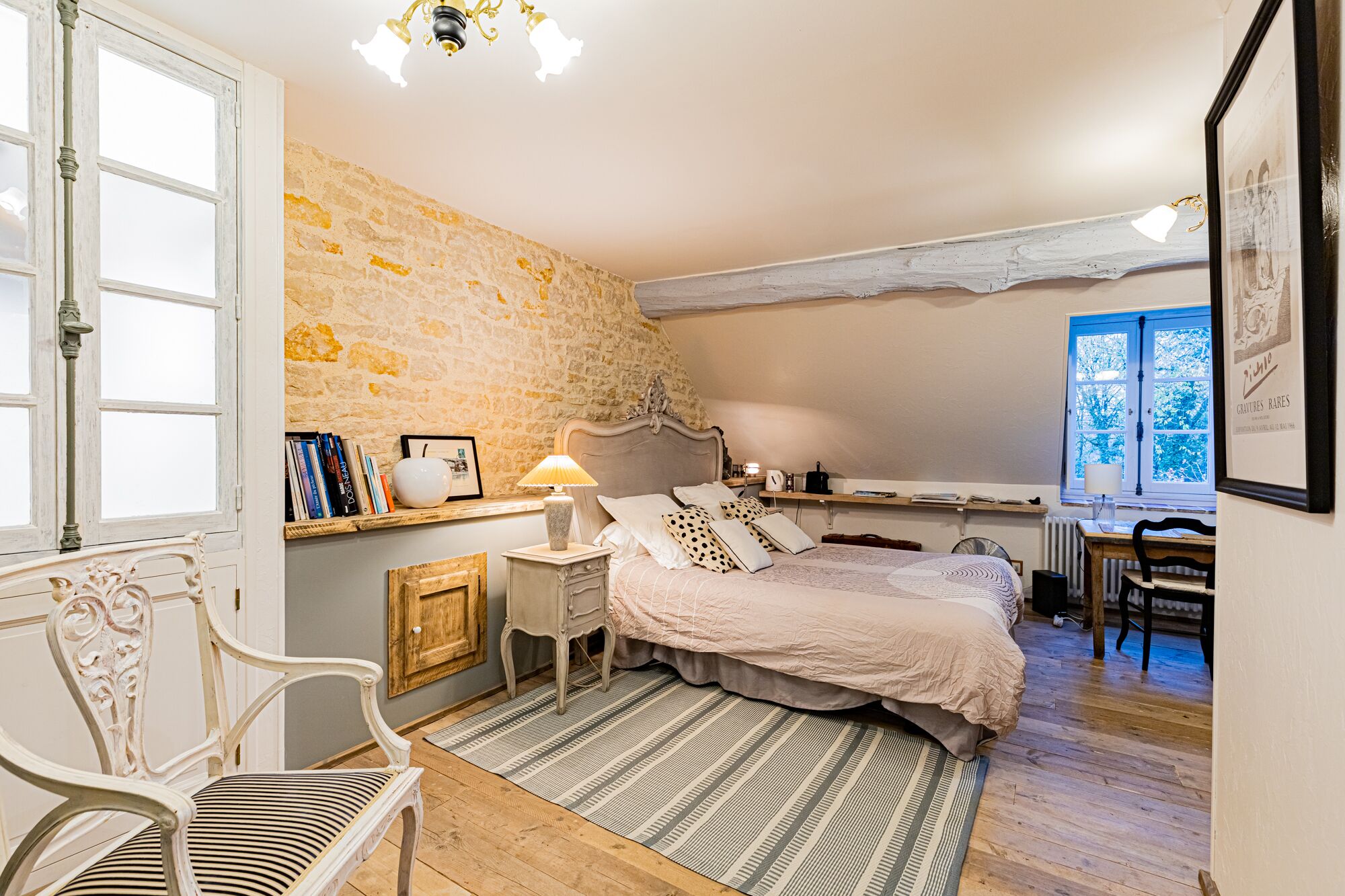 A bedroom with a stone wall, a bed with a headboard, a nightstand, a chair, a desk, and a window with a view.
