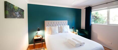 A neatly arranged bedroom with a large bed, a ceiling fan, a nightstand with a lamp, and a window with curtains.