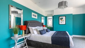 Un dormitorio moderno con paredes verde azulado, una cama con cabecera capitonada, una mesita de noche blanca, una lámpara roja y una araña.