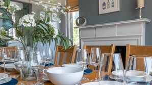 Un comedor con una mesa de madera dispuesta para una comida, un jarrón con flores blancas y una chimenea al fondo.