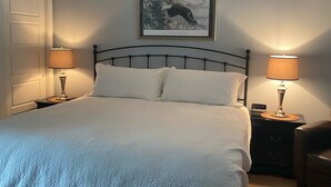 A neatly made bed with a white comforter and pillows, two bedside lamps, a framed picture of a bird on the wall, and a wooden floor. - Papa Bear Cottage (Downtown Cody) (Cody)