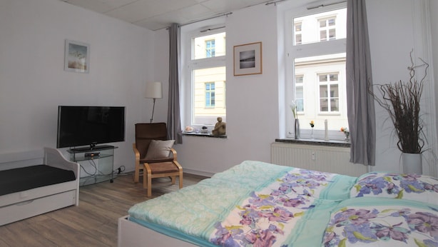 Habitación de hotel con cama, televisor, sillón, ventana con vistas a edificios y un florero con plantas.