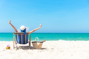 A person relaxing in a beach chair with arms raised, a straw drink, and a woven bag on a sandy beach.